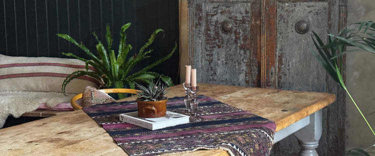 Pine table with a kilim runner, candlesticks and plants beside a distressed painted cupboard